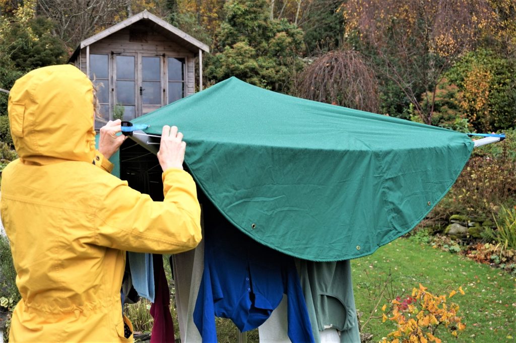 A woman wearing a yellow raincoat hangs laundry on a rotary dryer then covers it with a green Laundry Mac which protects the laundry from the rain